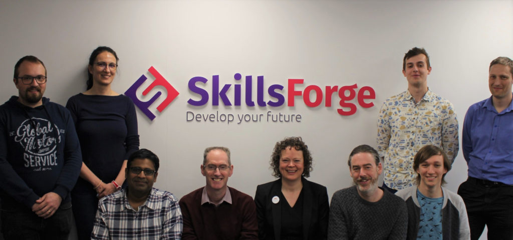 An image of the SkillsForge team, in their new offices, stood in front of the SkillsForge logo